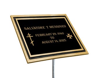 May include: A rectangular black plaque with gold-colored trim and text. The text reads "SALVATORE T. MESSINEO" above "FEBRUARY 29, 1940 TO AUGUST 14, 2025". There are two cross symbols and a Maltese cross symbol. The plaque is mounted on a silver rod.