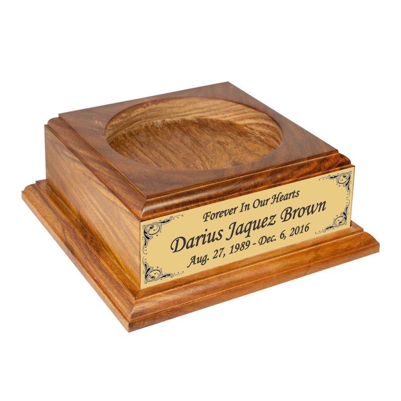 May include: A wooden urn with a square base and a circular opening. A gold-colored plaque on the front reads "Forever In Our Hearts" and the name "Darius Jaquez Brown" with dates. The wood has a warm, reddish-brown tone.