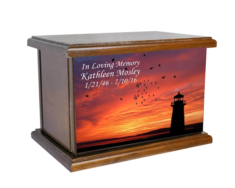May include: Wooden cremation urn featuring a sunset and lighthouse image. The urn is inscribed with the text "In Loving Memory Kathleen Mosley 1/21/46 - 7/10/16".