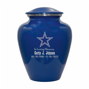 May include: A blue cremation urn with a silver lid. The urn has a white star with a silver outline and the text "In Loving Memory Curtis J. Johnson 09/26/1990-12/26/2022" engraved on the front.