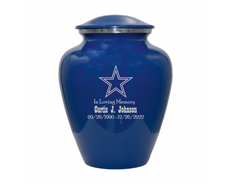 May include: A blue cremation urn with a silver lid. The urn has a white star with a silver outline and the text "In Loving Memory Curtis J. Johnson 09/26/1990-12/26/2022" engraved on the front.