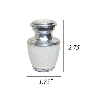 May include: Silver cremation urn with a white finish and engraved details. The urn is 2.75 inches tall and 1.75 inches wide.