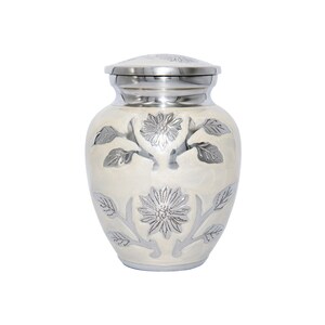 May include: A small white urn with a silver floral design. The urn has a lid and is made of metal.