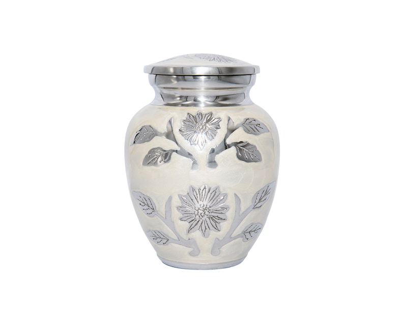 May include: A small white urn with a silver floral design. The urn has a lid and is made of metal.