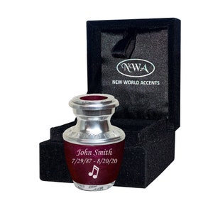 May include: A small silver urn with a burgundy lid and a personalized inscription that reads "John Smith 7/29/87-8/20/20" with a musical note symbol. The urn is in a black velvet box with the words "NWA NEW WORLD ACCENTS" printed on the lid.