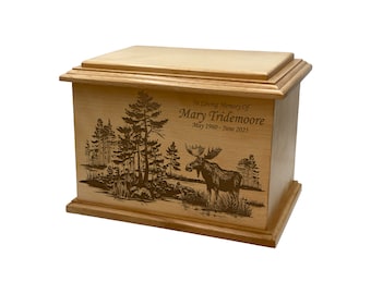 May include: A wooden urn with a light brown finish. The front features an engraved scene of a moose in a forest. The text "In Loving Memory of Mary Tridemore May 1960 - June 2023" is engraved on the front. The urn has a rectangular shape with a lid.
