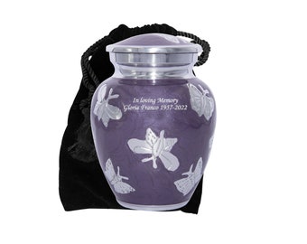 Puede incluir: Urna de cremación de color púrpura con diseño de mariposa plateada. La urna tiene una tapa plateada y está grabada con "In loving memory Gloria Franco 1957-2022".