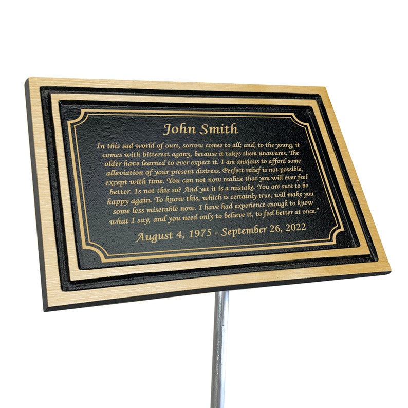 May include: A gold and black memorial plaque with the name John Smith and a quote about grief and sorrow. The dates August 4, 1975 - September 26, 2022 are also included.