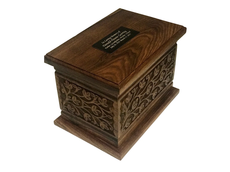May include: A dark brown wooden cremation urn with an engraved floral design and a brass plaque with the inscription "In loving memory of Donald Grace Sterling, Father, Grandfather, Friend, July 20, 1937 - March 11, 2021".