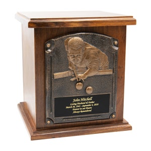 May include: A wooden cremation urn with a bronze plaque featuring a relief sculpture of a person playing pool. The plaque includes the name "John Hitchell", dates, and the text "Loving Husband & Father", "Forever in our Hearts", and "Always Remembered".