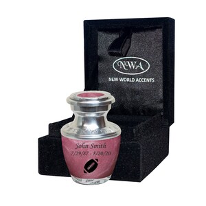 May include: A small, silver urn with a pink lid and a football design. The urn is engraved with the name "John Smith" and the dates "7/29/87 - 8/20/20". The urn is in a black velvet box with the words "NWA NEW WORLD ACCENTS" printed on the lid.