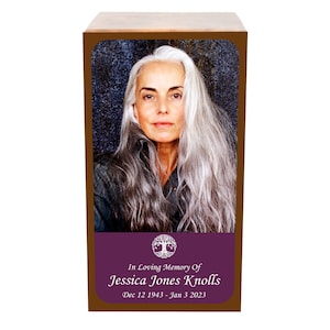 Könnte beinhalten: Eine Holzascheurne mit einem Foto einer Frau mit langen grauen Haaren. Die Urne hat ein violettes Band mit einem Lebensbaumsymbol und dem Text "In Loving Memory Of Jessica Jones Knolls 12. Dezember 1943 - 3. Januar 2023".