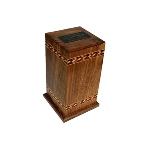 May include: A wooden cremation urn with a dark brown finish and intricate inlaid wood designs. The top of the urn has a small brass plaque with an inscription.