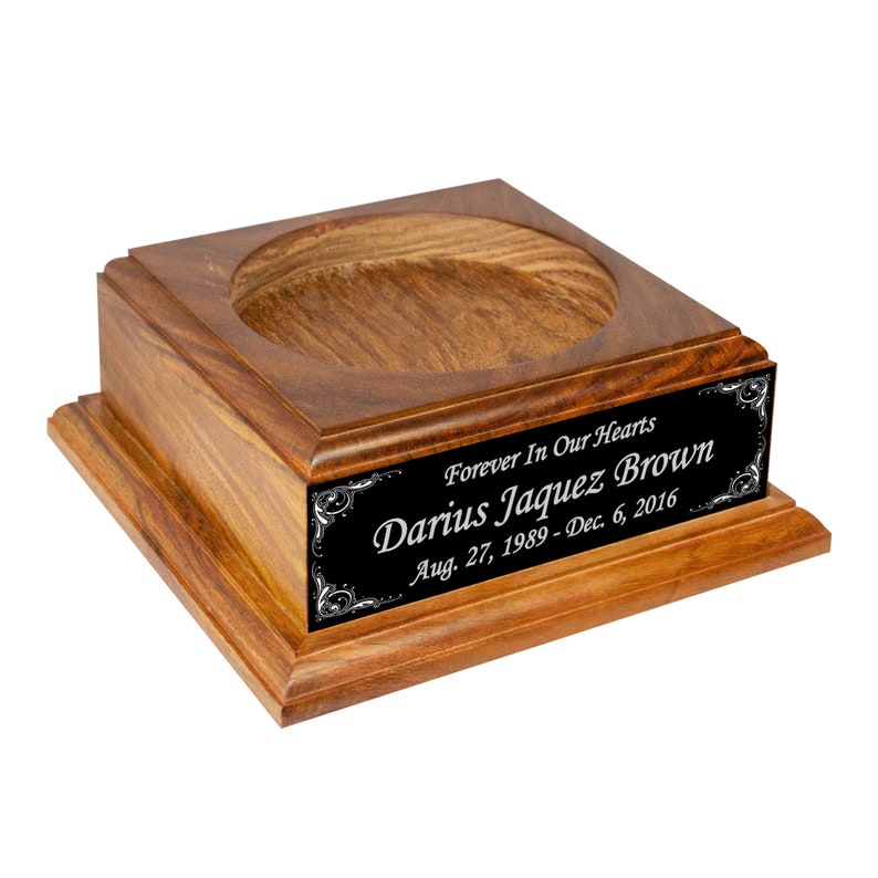 May include: A wooden urn with a square base and a circular opening at the top. A black plaque with white text reads "Forever In Our Hearts, Darius Jaquez Brown, Aug. 27, 1989-Dec. 6, 2016". The urn is made of a dark wood.