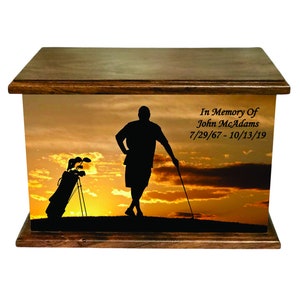 May include: Wooden cremation urn with a sunset scene featuring a golfer silhouette, golf bag, and clubs. The urn has a dark wood finish and includes the text "In Memory Of John McAdams 7/29/67 - 10/13/19".