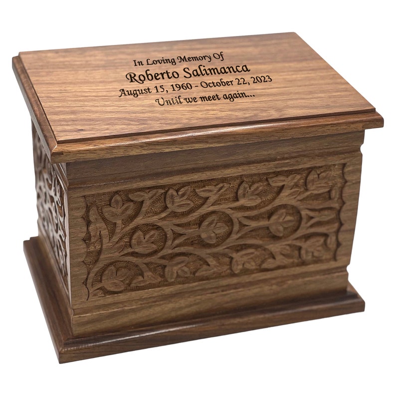 May include: A wooden cremation urn with an intricate carved floral design. The lid has an inscription that reads "In Loving Memory Of Roberto Salimanca August 15, 1960 - October 22, 2023 Until we meet again..."