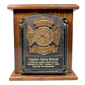 May include: A wooden cremation urn with a brass firefighter emblem. The emblem features a crossed axe and ladder with a fire helmet in the center. The urn has a black plaque with the inscription "Clayton Henry Brandt, Loving son, brother, uncle and dad, February 23, 1979 - October 27, 2020, Lake City Fire Department."