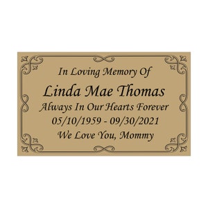 May include: A brown rectangular plaque with a decorative border and the text "In Loving Memory Of Linda Mae Thomas Always In Our Hearts Forever 05/10/1959 - 09/30/2021 We Love You, Mommy"
