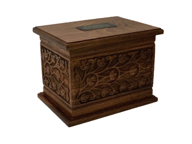 May include: A dark brown wooden box with an intricate carved floral design. The box has a flat top with a small brass plaque.