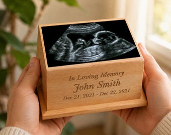 May include: A rectangular light-colored wooden box featuring a black and white ultrasound image on the top surface. The front panel is engraved with the text In Loving Memory, the name John Smith, and the dates Dec 21, 2021 - Dec 22, 2021.
