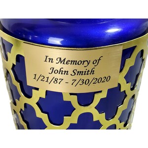 May include: A blue cremation urn with a gold metal lattice design and a gold plaque that reads "In Memory of John Smith 1/21/87 - 7/30/2020".