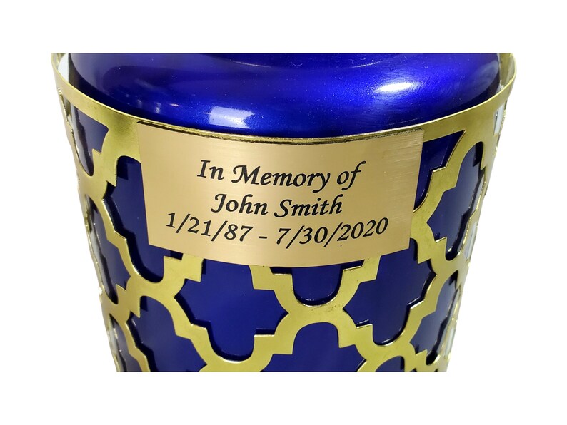 May include: A blue cremation urn with a gold metal lattice design and a gold plaque that reads "In Memory of John Smith 1/21/87 - 7/30/2020".