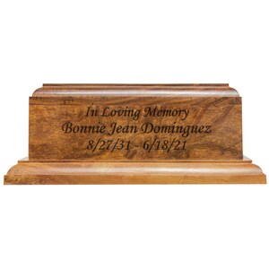 May include: A wooden urn with a rectangular shape and a warm brown finish. The front of the urn is engraved with the words "In Loving Memory," the name "Bonnie Jean Dominguez," and the dates 8/27/31 - 6/18/21.