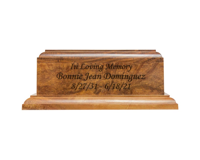 May include: A wooden urn with a rectangular shape and a warm brown finish. The front of the urn is engraved with the words "In Loving Memory," the name "Bonnie Jean Dominguez," and the dates 8/27/31 - 6/18/21.