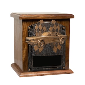 May include: A wooden urn with a bronze plaque featuring a vintage car and chequered flags. The plaque has a blank space for engraving.