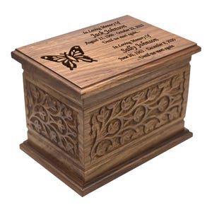May include: A wooden cremation urn with an intricate carved floral design. The lid features a carved butterfly and personalized text: "In loving memory of Jack Johnson August 15, 1960-October 22, 2023 Until we meet again In loving memory of Sally Johnson June 20, 1963-December 8, 2020 Until we meet again"