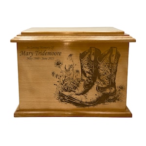 May include: A wooden urn with a light brown finish. The front features an engraved design of cowboy boots and wildflowers. Text reads "In Loving Memory of Mary Tridemoore May 1960 - June 2023". The urn has a rectangular shape with a lid.