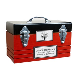 May include: A red wooden toolbox with black trim and silver latches. The toolbox has a black and white label with the text "In Living Memory" and a graphic of tools. The label below it reads "James Robertson 9/27/1945 - 9/27/2021 Forever In Our Hearts".