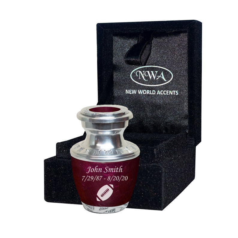 May include: A small, silver and burgundy urn with a football design. The urn is engraved with the name "John Smith" and the dates "7/29/87 - 8/20/20". The urn is in a black velvet box with the words "NWA NEW WORLD ACCENTS" on the lid.