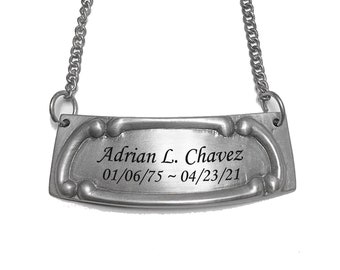 Puede incluir: Etiqueta de botella de vino de metal plateado con una cadena. La etiqueta tiene el nombre "Adrian L. Chavez" y las fechas "01/06/75 ~ 04/23/21" grabadas. La etiqueta tiene un diseño curvo y ornamentado.