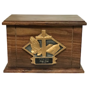 May include: A wooden cremation urn with a gold-toned cross, praying hands, and an open book design. The urn has a black plaque with the text "In loving memory Jorge Jung 2015-2022".