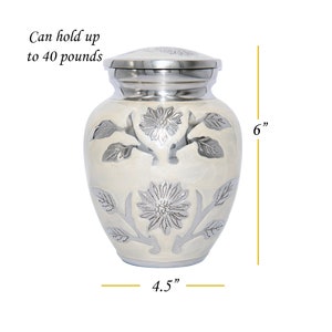 May include: White cremation urn with a silver floral design. The urn is 6 inches tall and 4.5 inches wide. It can hold up to 40 pounds.