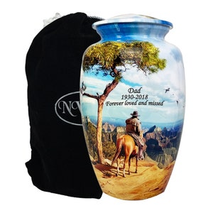 Puede incluir: Urna funeraria azul y blanca con diseño de paisaje. La imagen presenta un vaquero a caballo, un árbol y el texto "Dad 1930-2018 Forever loved and missed". Se incluye una bolsa de terciopelo negro.