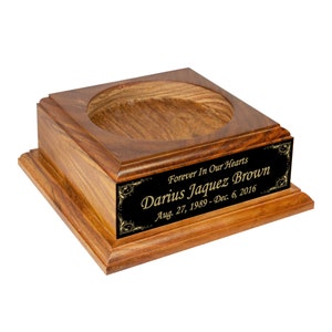 May include: A wooden urn with a square base and a circular opening at the top. A black plaque with gold lettering reads "Forever In Our Hearts, Darius Jaquez Brown, Aug. 27, 1989-Dec. 6, 2016."