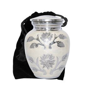 May include: A white cremation urn with a silver floral design. The urn has a lid and comes with a black velvet bag.