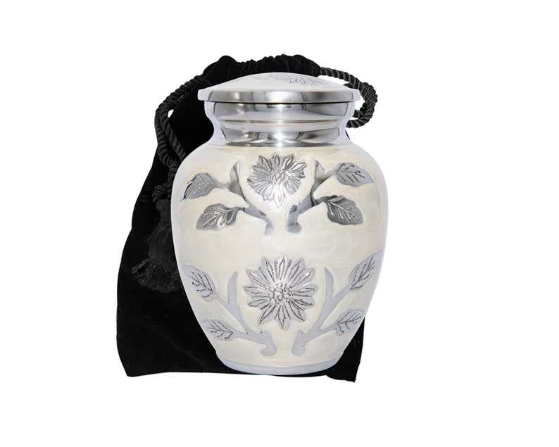 May include: A white cremation urn with a silver floral design. The urn has a lid and comes with a black velvet bag.