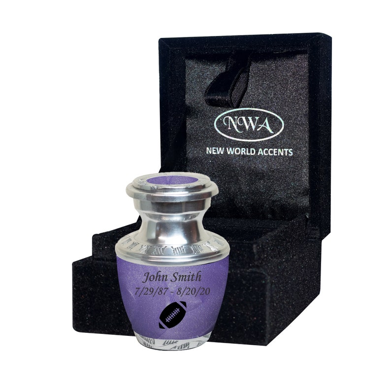 May include: A small silver urn with a purple lid and a football design. The urn is engraved with the name "John Smith" and the dates "7/29/87 - 8/20/20". The urn is in a black velvet box with the words "NWA NEW WORLD ACCENTS" printed on the lid.