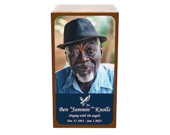 May include: A wooden memorial urn with a framed portrait of an older Black man wearing a hat and blue striped shirt. The image includes text that reads "Ben 'Jammin' Knolls, Singing with the angels, Dec 12 1943 - Jan 3 2023."