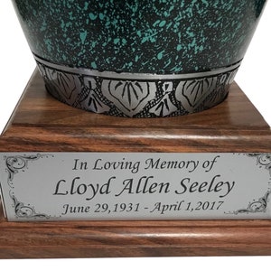May include: A teal and black urn with a decorative silver band sits on a wooden base. A silver plaque reads "In Loving Memory of Lloyd Allen Seeley June 29, 1931 - April 1, 2017."