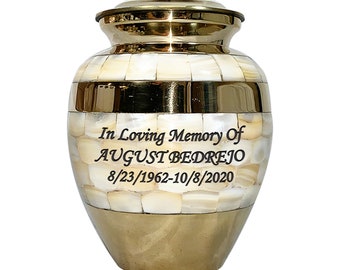May include: A decorative urn with a gold-colored base, lid, and bands. The body is made of cream-colored, mosaic-style tiles. The text "In Loving Memory Of AUGUST BEDREJO 8/23/1962-10/8/2020" is printed on the front.