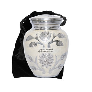 May include: A small white cremation urn with a silver floral design and engraved inscription. The urn is in a black velvet bag with a drawstring.