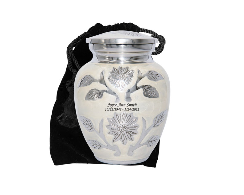 May include: A small white cremation urn with a silver floral design and engraved inscription. The urn is in a black velvet bag with a drawstring.