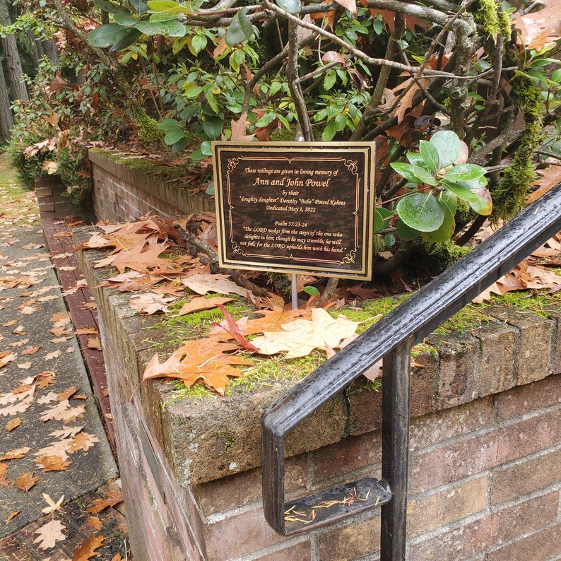 May include: A gold plaque with a black background is mounted on a wooden post. The plaque reads "These plantings are given in loving memory of Ann and John Powell by their lovingly daughter "Serenity" Hale" Powell-Kiddens Dedicated May 1, 2022 Psalm 17:23-24 "The LORD makes firm the steps of the one who delights in him; though he may stumble, he will not fall, for the LORD upholds him with his hand."