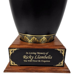 May include: A black urn with a gold decorative band and a wooden base. The urn has a black plaque with gold text that reads "In Loving Memory of Ricky Lambelis, You Will Never Be Forgotten."