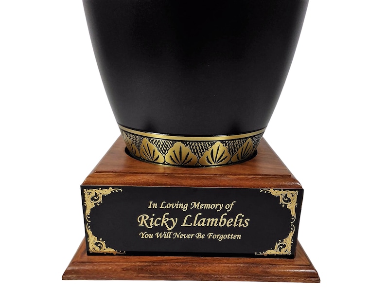 May include: A black urn with a gold decorative band and a wooden base. The urn has a black plaque with gold text that reads "In Loving Memory of Ricky Lambelis, You Will Never Be Forgotten."