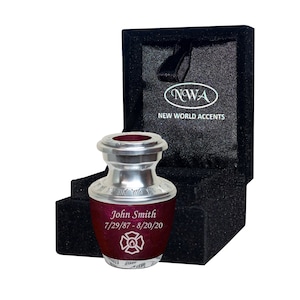 May include: A burgundy and silver cremation urn, engraved with "John Smith" and the dates 7/29/87 - 8/20/20, and a firefighter emblem. It's displayed in a black velvet box, branded with "NWA NEW WORLD ACCENTS".